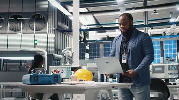 Black solar panel engineer in a high tech facility doing an inspection, looking at efficiency and production line metrics on laptop. Expert reviewing operations for sustainability. Camera B. video
