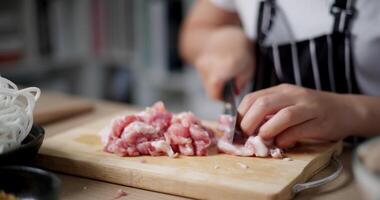 Handheld selective focus shot, Asian woman's hands cooking by slicing pork on a wooden cutting board to make noodles, preparing food. video