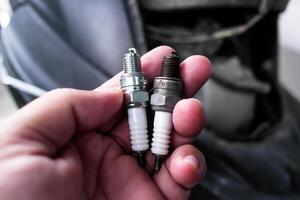 Spark Plug, A hand holds two spark plugs, one new and one used, against a blurred automotive background, highlighting the difference in wear. photo