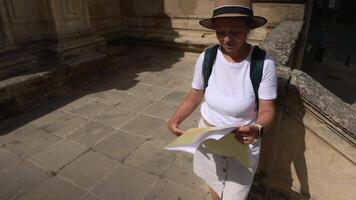 Joyful woman tourist in a hat and backpack explores an old medieval city. She smiles while navigating historic streets with a map, discovering a new travel destination on a bright sunny day. video