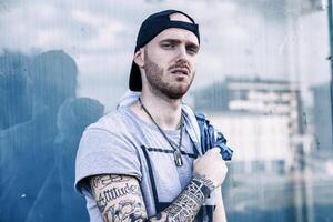 portrait of young tattooed rapper looking into camera in front of large window in the outskirts photo