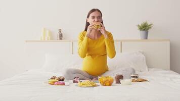 A pregnant woman sits cross-legged on a bed, joyfully holding a burger. Various snacks like fries, donuts, and cookies are spread around. The setting is bright and cozy. video
