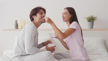 A joyful couple shares a meal, seated comfortably on their bed in a serene indoor setting. They smile and engage in playful banter, creating a warm atmosphere. video