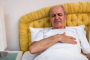 Mature Caucasian man with gray waking up in bed, holding his chest with distressed expression due to heart pressure. photo