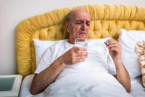Worried elderly man with gray hair drinking pills while lying awake in bed in the morning, looking anxious and tired, showing aging process and health struggles. photo