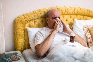 Sick and exhausted senior man having flu and sneezing into tissue while lying on bed surrounded by medication, represents respiratory issues, cold or allergies. photo