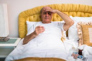 Old man lying in bed, checking his temperature with thermometer, suffering from flu symptoms at home. photo