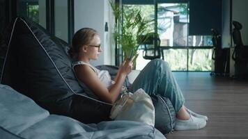 Young woman employee is taking break from work, seated on beanbag chair in modern office space, browsing her smartphone. Procrastination and departure from reality on Internet via smartphone video