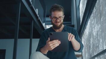 Happy Digital Marketer with beard and glasses using digital tablet while sitting in modern office, enjoying his work and using technology for business. Content Creator creates new podcast video