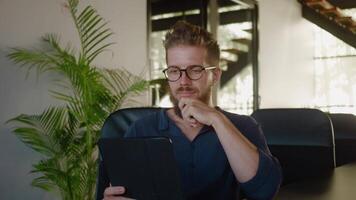 Data analyst with eyeglasses is sitting in chair and reading information on his tablet, touching his chin with, in modern office environment. Concept of data analysis and profit in business video