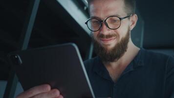 Project Manager working from home at night, holding tablet computer and smiling, enjoying comfort of his cozy space while staying connected and productive. Business Analyst views successful quotes video