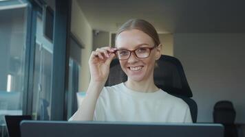 Product manager with eyeglasses smiles while adjusting her glasses and working on her laptop in modern office environment, exuding confidence and professionalism. Product manager Successful woman video