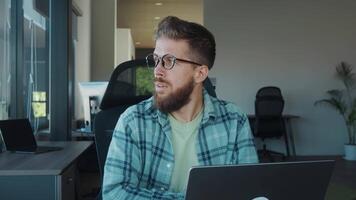 jovem empreendedor com barba e Óculos concentrando em dele trabalhos em computador portátil dentro moderno casa escritório, demonstrando flexibilidade do controlo remoto trabalhar. conceito do trabalhos dentro coworking video