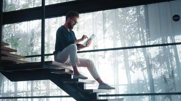 Businessman is using digital tablet while sitting on stairs in modern office with large windows and bamboo shadows, representing concepts of technology, business, and modern workspaces video