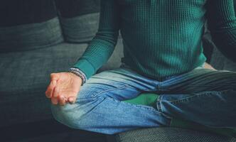 Dressed in casual clothes and stone bead bracelets, a man in his 55s practices meditation on his couch. The soft natural light and calm posture reflect a grounded, mindful lifestyle in a comfortable photo