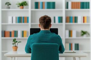 A man sits at a desk in a home office, working on his computer. The room is modern and minimalist, with bookshelves filled with bo photo