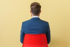 A man in a suit sits on a red chair, viewed from behind. The image is simple yet evocative, capturing a moment of contemplation or photo