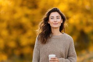 Smiling woman enjoying a coffee in an autumn park. A beautiful portrait of a happy person surrounded by golden leaves and warm su photo