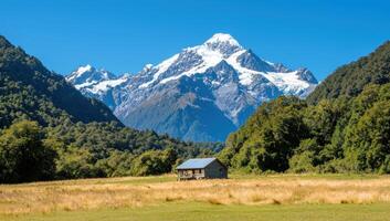 Rural hut sits nestled below peak with mountains and trees in background photo