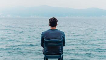 Lonely adult man sitting alone by the sea at sunset looking away, back view. photo