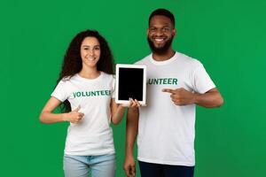 Diverse Volunteers Holding Digital Tablet Pointing At Blank Computer Screen And Gesturing Thumbs Up Standing In Studio On Green Background, Smiling To Camera. Mockup photo