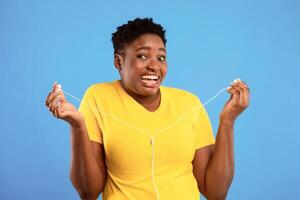 Auwful Playlist. Funny Black Lady Holding Earphones Not Liking Music Standing Over Blue Studio Background, Looking At Camera In Disgust. I Don't Want To Hear It Concept photo
