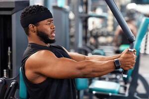 Side view of black guy bodybuilder making work out for shoulders and chest muscles at gym, copy space. Concentrated african american man exercising on pec deck machine, closeup photo