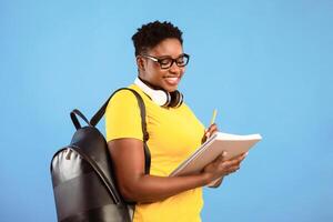 Black Plus-Size Student Lady Learning Posing Holding Books Standing Over Blue Studio Background, Wearing Eyeglasses And Taking Notes. Knowledge, College And University Study Concept photo
