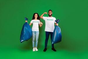 Two Diverse Volunteers Holding Blue Garbage Bags Showing Biceps And Pointing At Volunteer Inscription On T-Shirts Posing Over Green Studio Background. Keep Environment Clean. Full Length photo