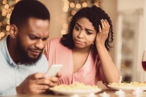 Bad Date. Unhappy Sad African American Woman Is Getting Bored Sitting On Date In Restaurant While Her Boyfriend Using Mobile Phone And Chatting, Ignoring His Girlfriend. Relationship Problem photo