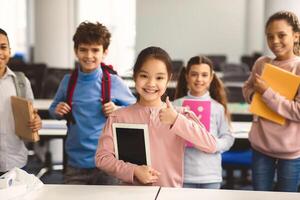 Education And Technology Concept. Smiling small asian girl holding digital tablet and showing thumbs up sign gesture. Diverse group of international classmates standing with backpacks in background photo