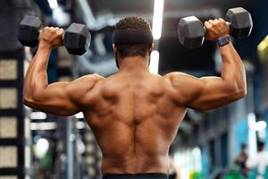 Back view of african american shirtless man bodybuilder with muscular body exercising at modern gym, lifting up dumbbells, black man making work out for his biceps and back, copy space photo