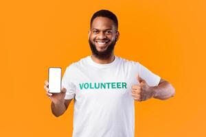 Happy Black Volunteer Guy Showing Empty Phone Screen And Gesturing Thumbs Up Standing On Yellow Studio Background. Mobile Application For Charity And Volunteering. Mockup photo
