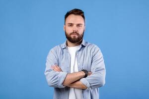 Portrait of serious millennial guy posing with crossed arms and looking at camera on blue studio background. Confident bearded young man in casual clothes having cool face expression photo