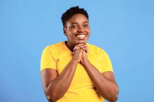 Please Concept. Overweight African American Lady With Clasped Hands Begging Making Pleading Gesture Smiling Looking At Camera Posing Standing Over Blue Background. Studio Shot. photo