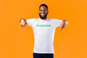 African Volunteer Guy Pointing Fingers Down Posing Smiling To Camera Over Yellow Background. Activist Advertising Something Standing In Studio. Volunteering Advertisement. Look There Concept photo