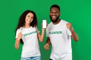 Two Mixed Volunteers Showing Mobile Phone Blank Screen Gesturing Yes In Joy Recommending Volunteering Application Standing On Green Background. Studio Shot. Call Us Concept. Mockup photo
