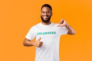 I like volunteering. Smiling african volunteer man pointing thumb at himself gesturing thumbs-up posing on yellow studio background, smiling to camera. Activist's portrait photo