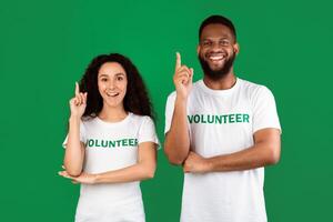 Two Multiracial Volunteers Having Idea Pointing Fingers Up Posing Standing Over Green Studio Background, Smiling To Camera. Volunteering Work Concept. Activists Have Eureka Moment photo