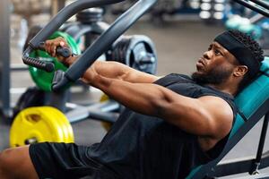Athletic young black man having workout on chest press machine in gym, pushing weight while reclining on bench, building up arms and torso muscles, side view, panorama, closeup photo
