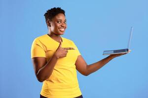 Cheerful African Woman Holding Laptop Pointing Finger At Computer Advertising Website Standing Over Blue Background, Smiling To Camera. Black Lady Posing With Laptop In Studio photo