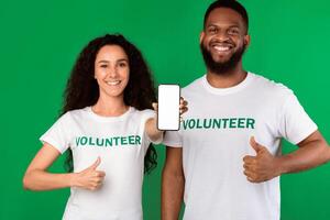 Two Cheerful Volunteers Showing Phone Empty Screen Gesturing Like Approving Charity Application Standing On Green Studio Background, Smiling To Camera. Volunteering Work, Call Us Concept. Mockup photo