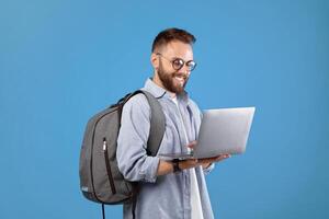 Cool millennial guy with backpack using laptop for online work or studies over blue studio background. Happy young man communicating on web, watching movie, browsing internet photo
