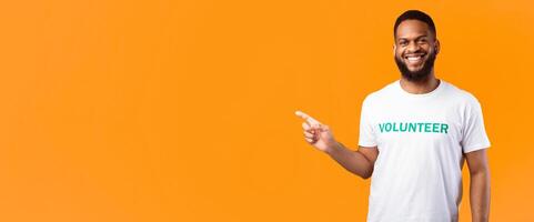 Black Male Volunteer Pointing Finger Aside Smiling To Camera Standing Over Yellow Studio Background. Activist Advertising Volunteering Project. Look There Concept. Panorama, Copy Space photo