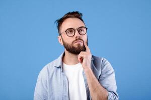 Portrait of pensive bearded man in glasses touching his chin, deep in thought over blue studio background. Puzzled young guy making choice or decision, dreaming about something photo