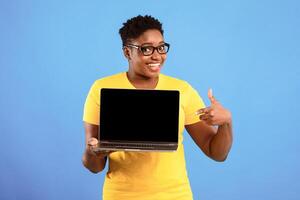 Happy African American Lady Showing Laptop Empty Screen Standing Over Blue Studio Background. Female Holding Computer With Black Monitor, Recommending Online Website Concept. Mockup photo