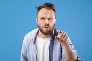 It is your fault. Angry Caucasian man pointing finger at screen in accusation on blue studio background. Frustrated young guy blaming you. Negative human emotions concept photo