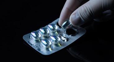 Hand in glove taking shimmering iridescent capsule from blister pack against dark background photo