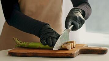 A chef in gloves skillfully cuts fresh asparagus on a wooden chopping board. The kitchen is bright and eco-friendly, emphasizing healthy cooking practices. Attention to detail is evident. video