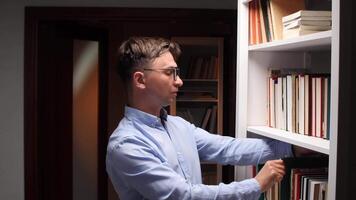 Young man searching intently for a specific book among the organized shelves of a home library, surrounded by a diverse collection of literature and knowledge video